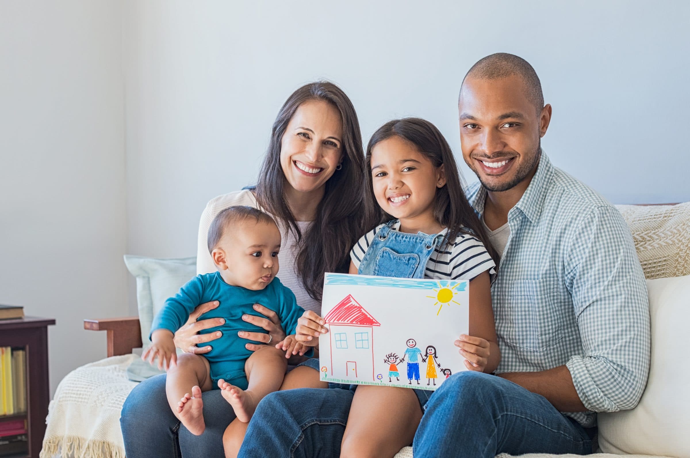 Daughter showing drawing of a happy family in new house. Cute infant looking at colorful drawing of his sister. Happy proud multiethnic parents sitting with children on sofa  and looking at camera.